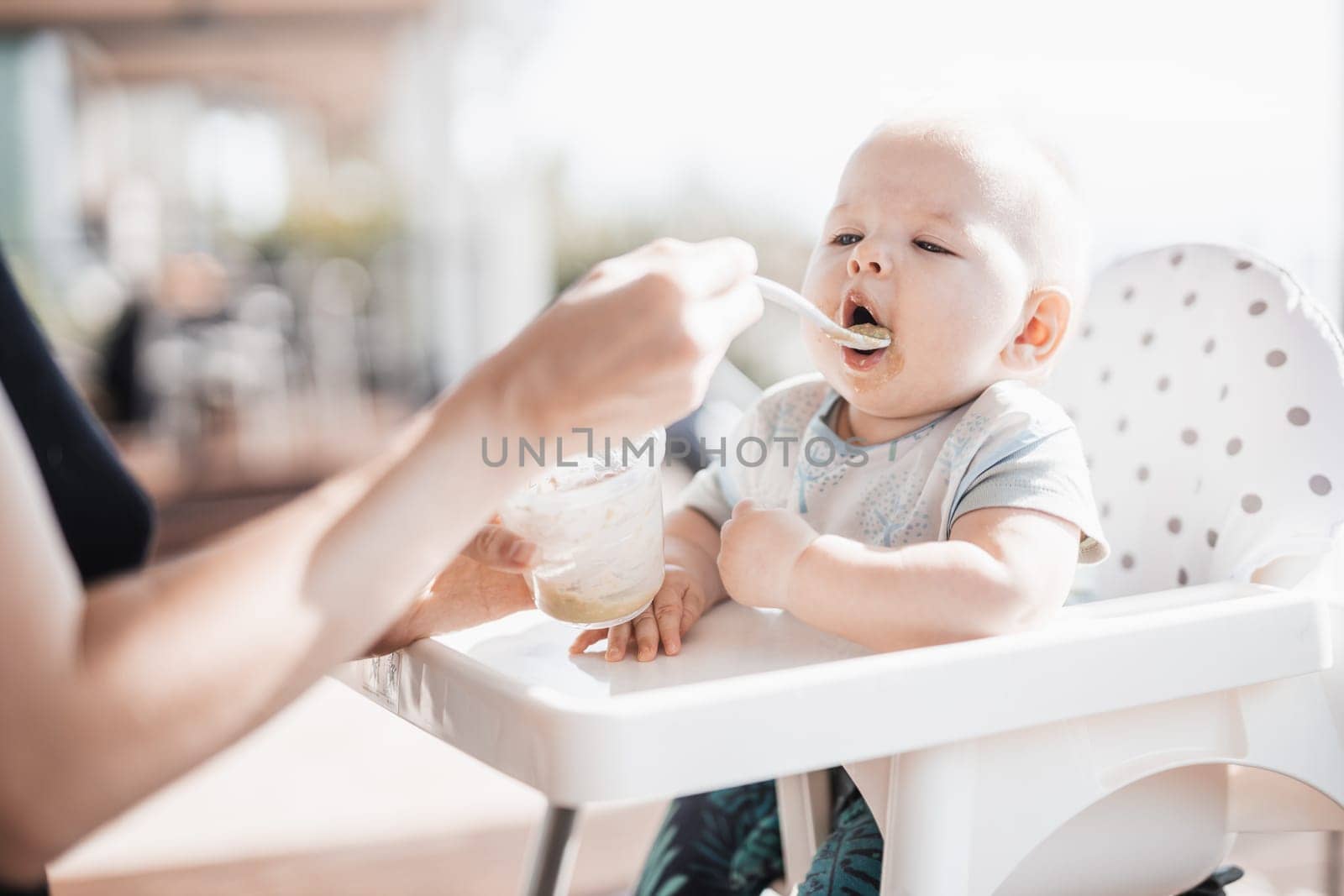 Mother spoon feeding her baby boy child in baby chair with fruit puree on a porch on summer vacations. Baby solid food introduction concept