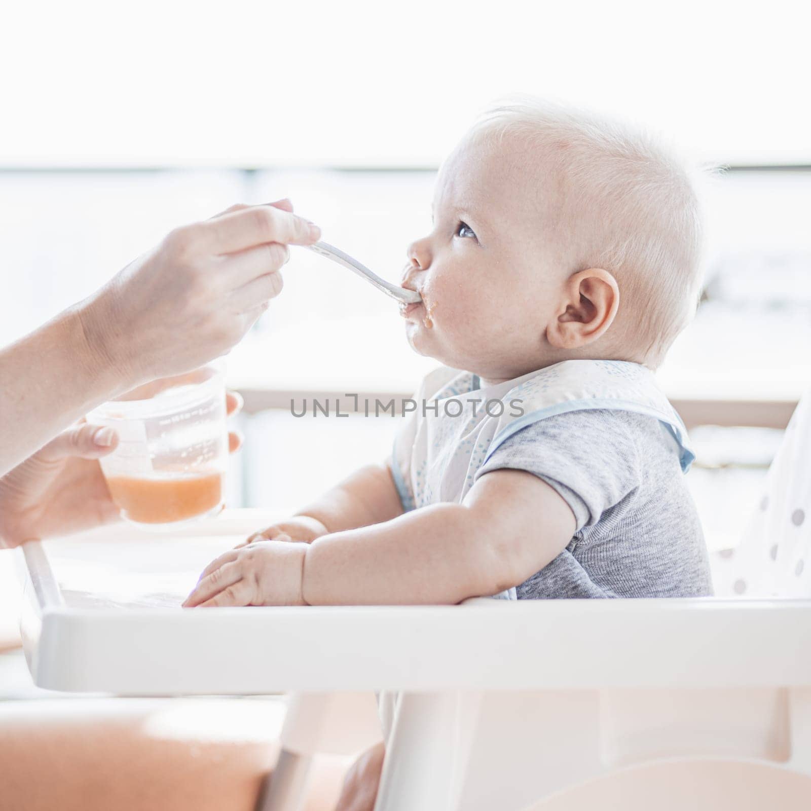 Mother spoon feeding her baby boy child in baby chair with fruit puree on a porch on summer vacations. Baby solid food introduction concept. by kasto