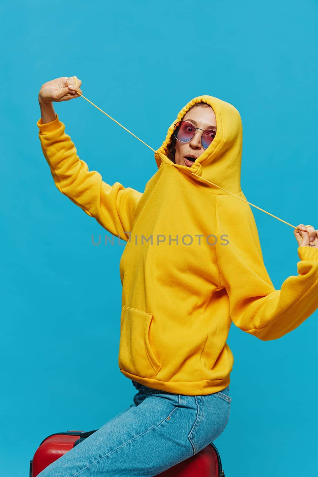 Smile woman sitting on a suitcase in a yellow hoodie, blue jeans and glasses on a blue background, packing for a trip. High quality photo