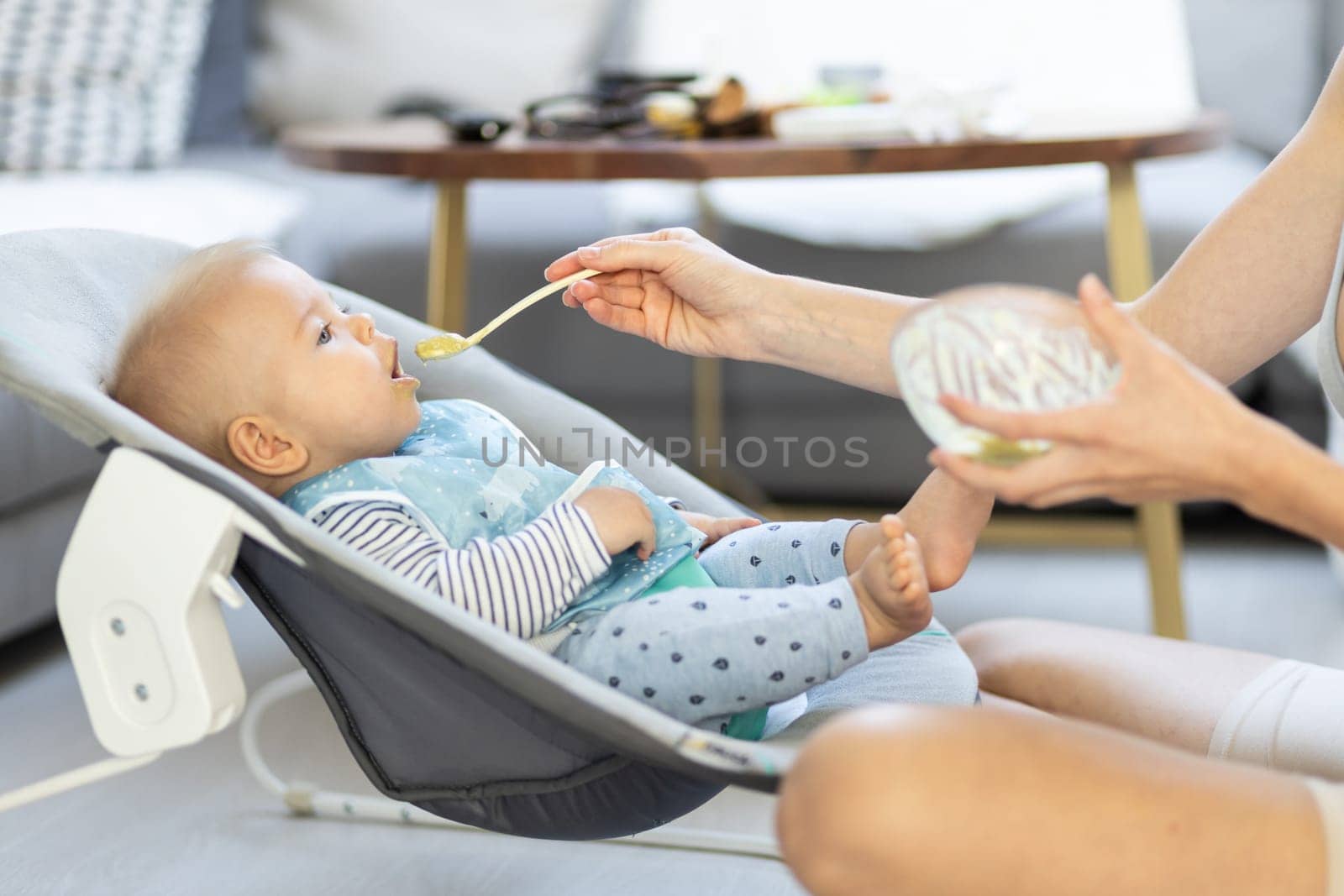 Mother spoon feeding her baby boy infant child in baby chair with fruit puree. Baby solid food introduction concept. by kasto