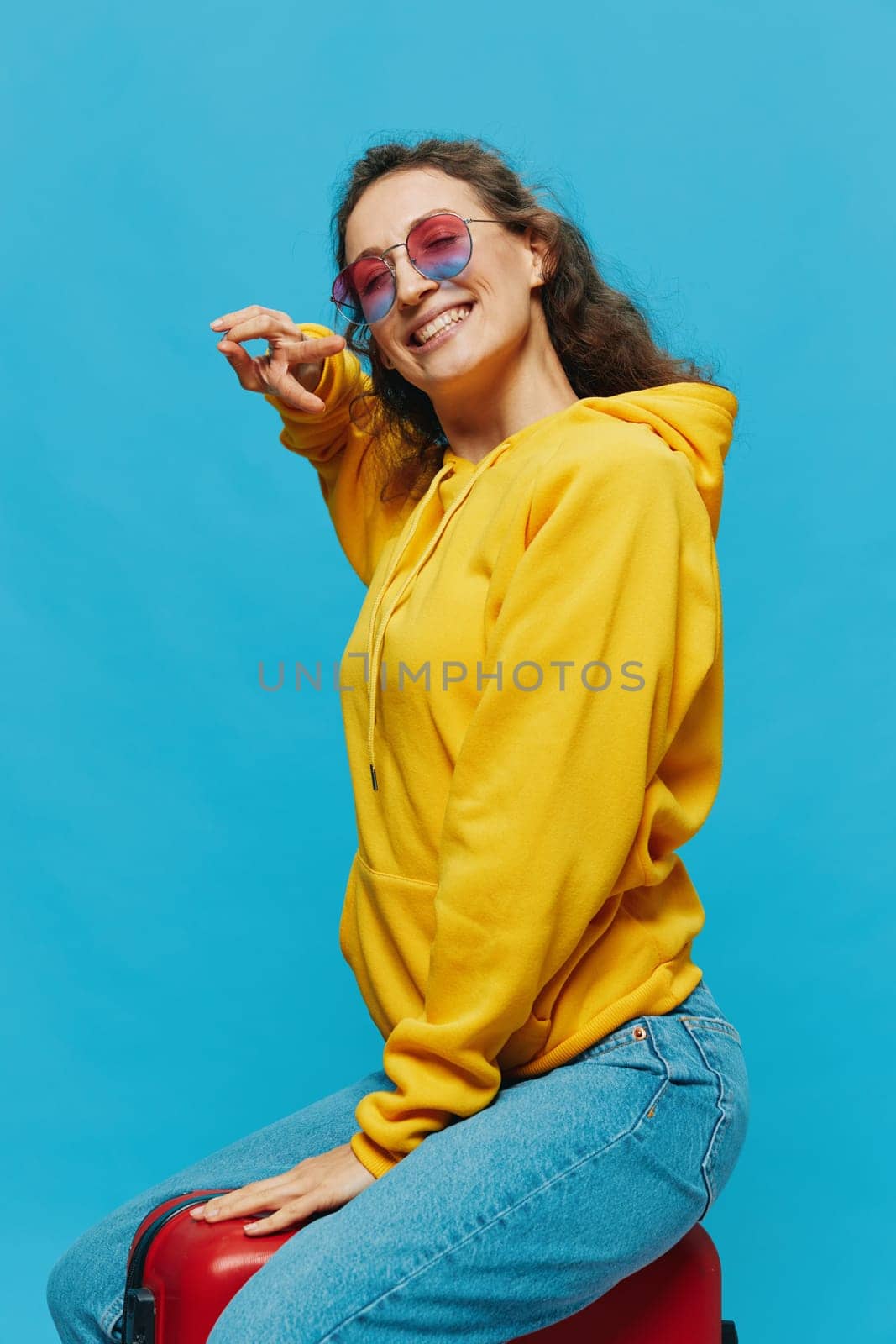 Smile woman sitting on a suitcase in a yellow hoodie, blue jeans and glasses on a blue background, packing for a trip. High quality photo