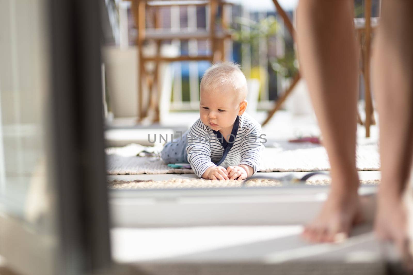 Cute little infant baby boy playing with toys outdoors at the patio in summer being supervised by her mother seen in the background. by kasto