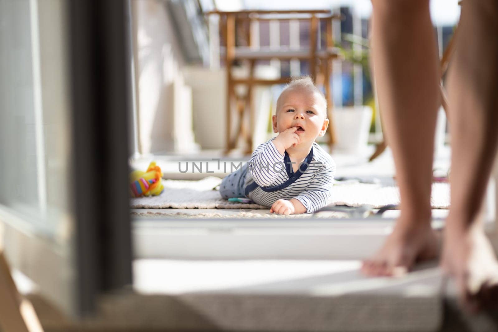 Cute little infant baby boy playing with toys outdoors at the patio in summer being supervised by her mother seen in the background. Selective focus.