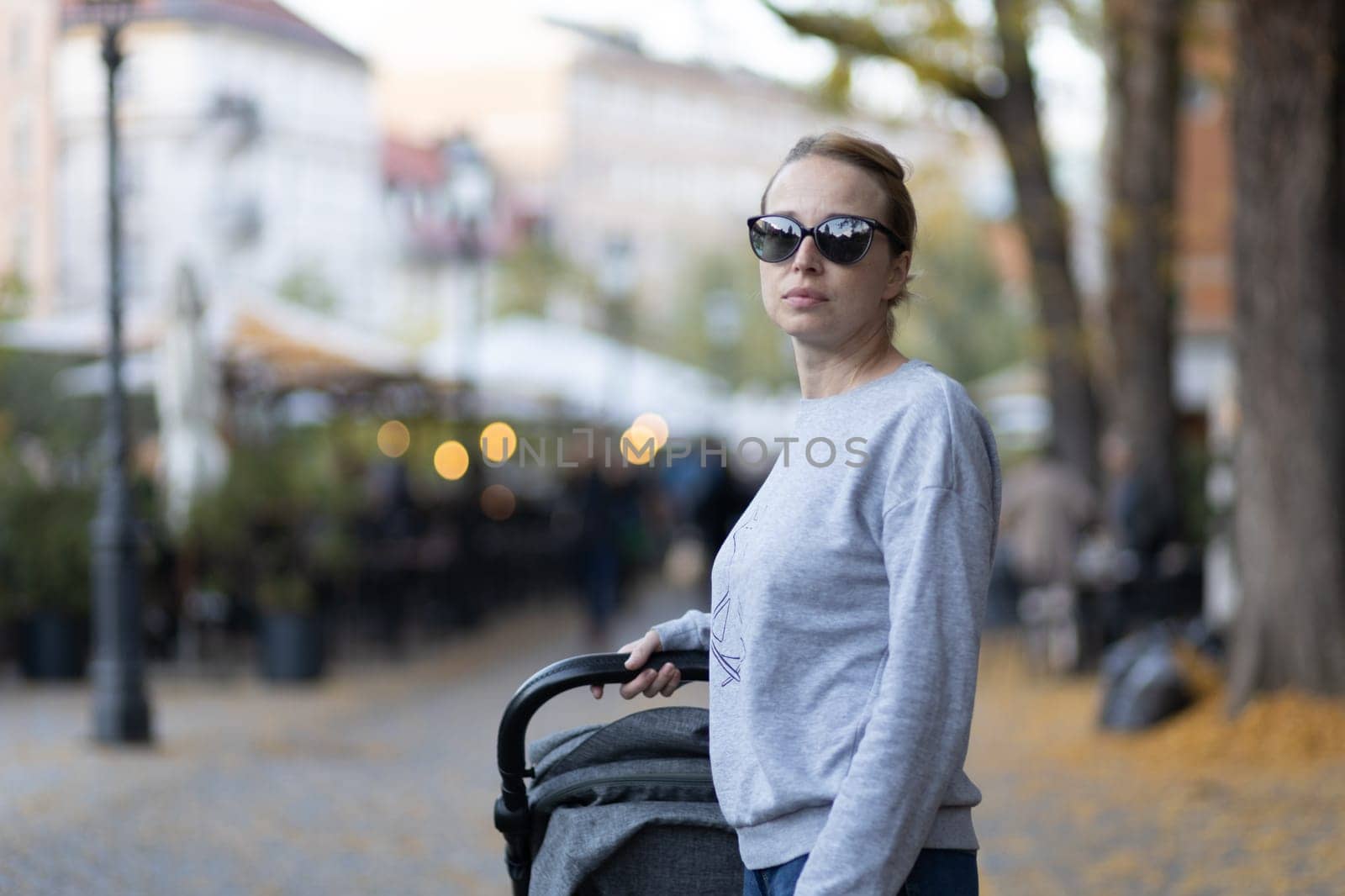 Stylish young woman wearing sunglasses walking through Ljubljana city center pushing and rocking baby stroller. Warm autumn or spring weather for outdoor activity. by kasto