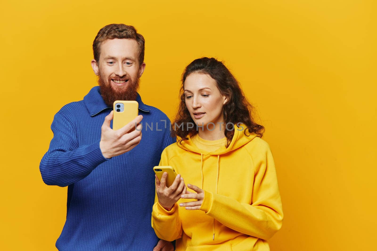 Man and woman couple smiling merrily with phone in hand social media viewing photos and videos, on yellow background, symbols signs and hand gestures, family freelancers. by SHOTPRIME