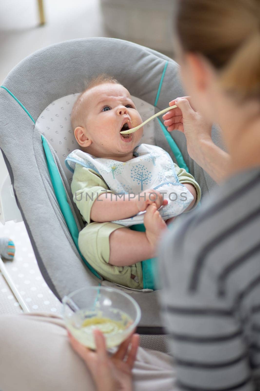 Mother spoon feeding her baby boy child in baby chair with fruit puree. Baby solid food introduction concept