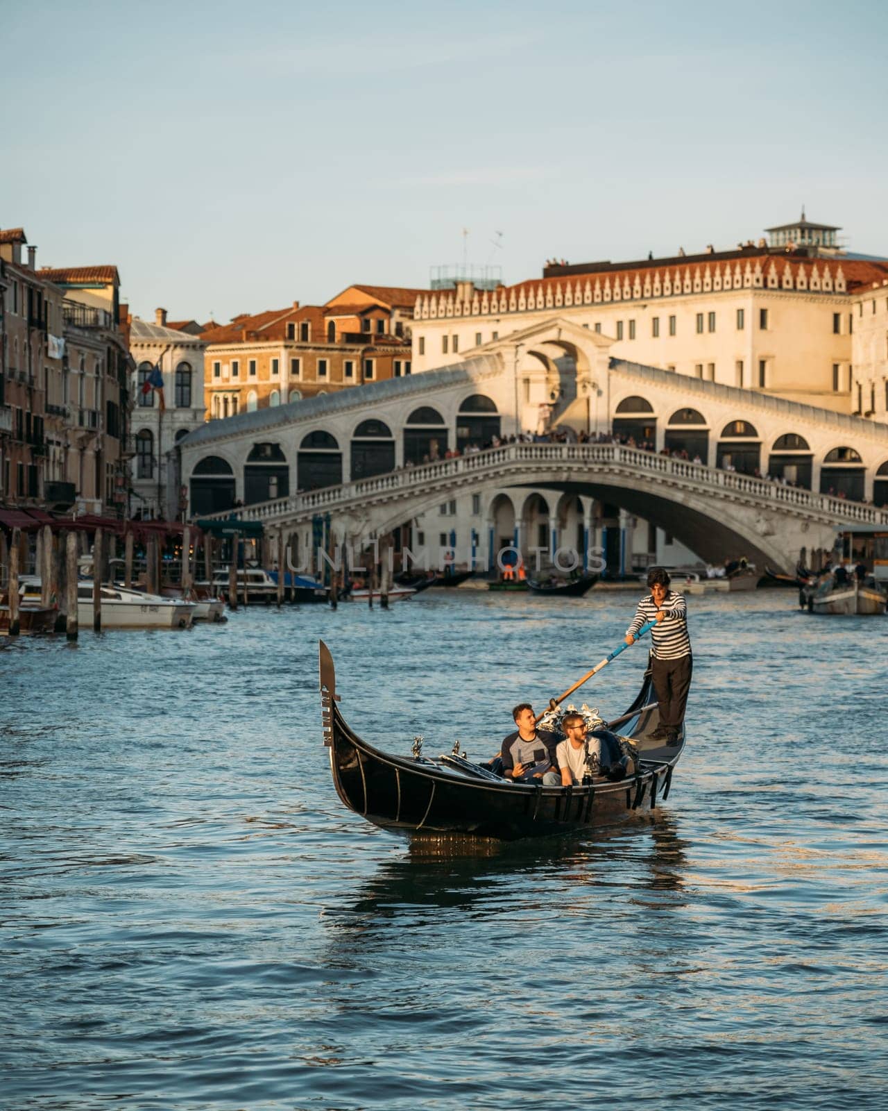 Beautiful view of tourists sitting in gondola with a gondolier, floating along canal on the backdrop of Rialto bridge on sunny warm summer evening. Concept of tourism in the romantic city of Italy by apavlin
