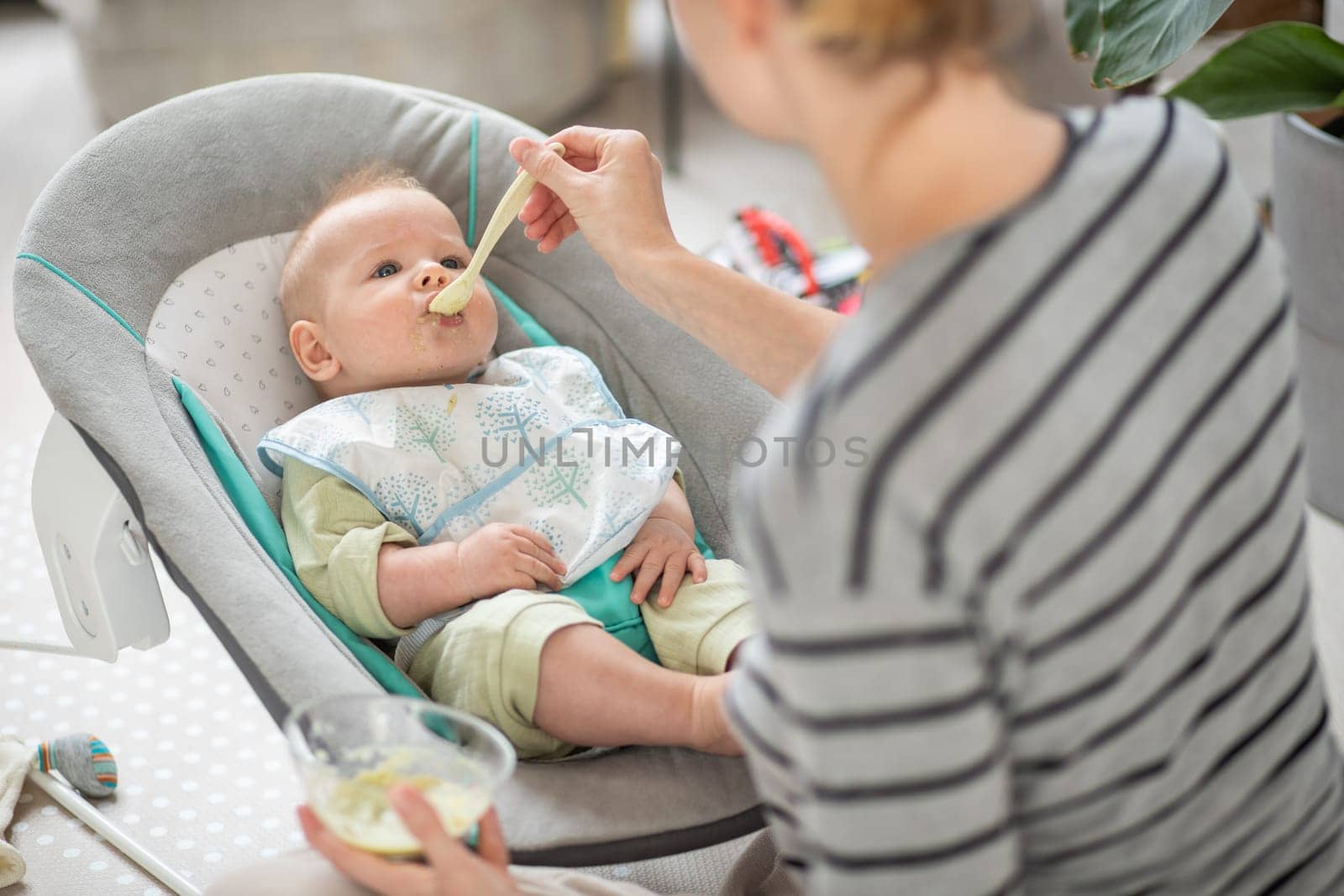 Mother spoon feeding her baby boy child in baby chair with fruit puree. Baby solid food introduction concept