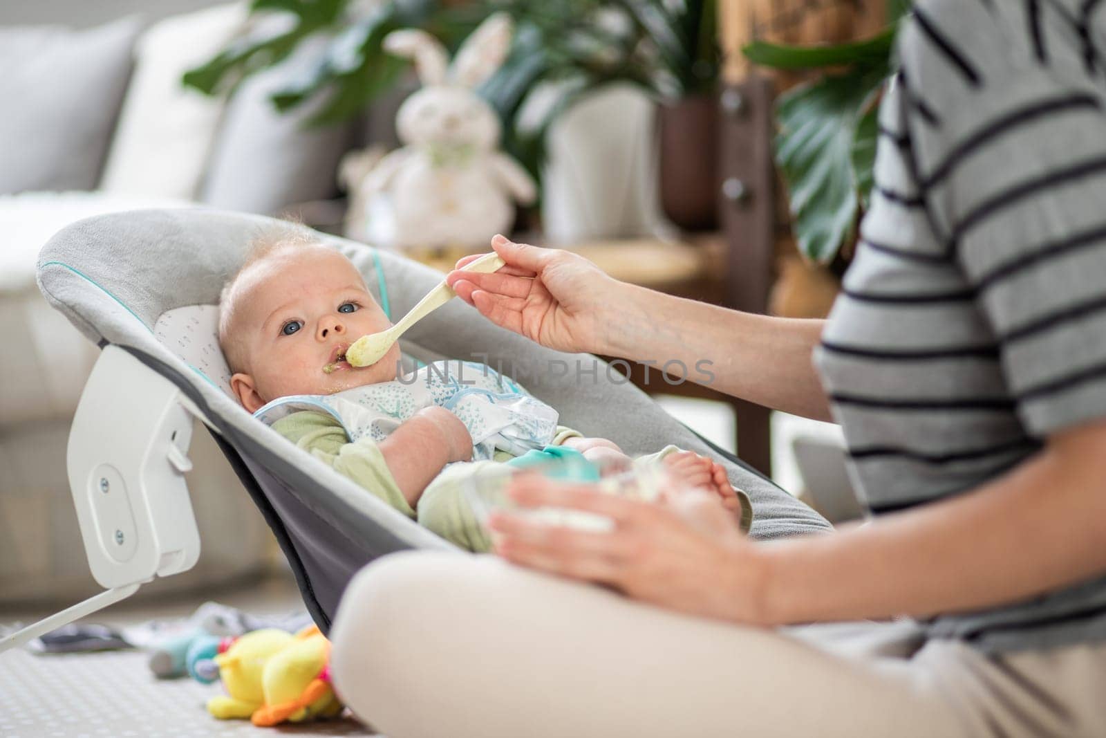 Mother spoon feeding her baby boy infant child in baby chair with fruit puree. Baby solid food introduction concept. by kasto