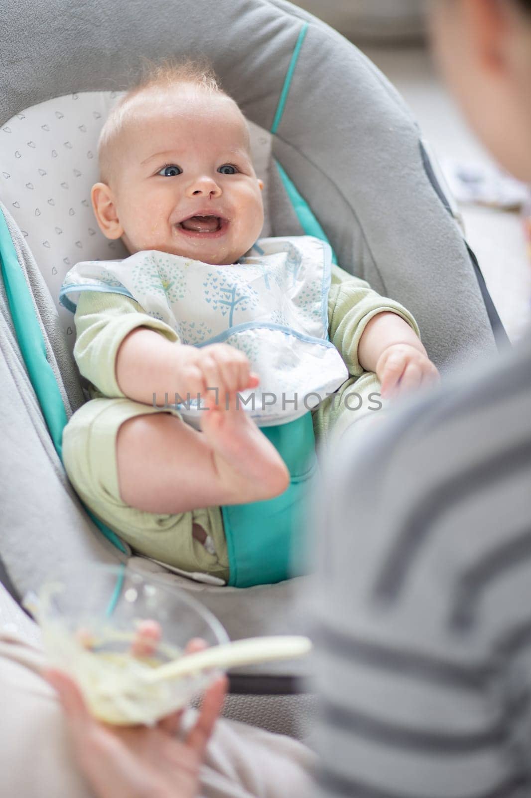 Mother spoon feeding her baby boy child in baby chair with fruit puree. Baby solid food introduction concept