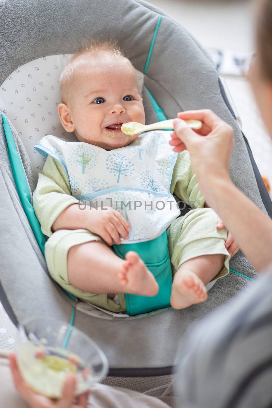 Mother spoon feeding her baby boy infant child in baby chair with fruit puree. Baby solid food introduction concept. by kasto