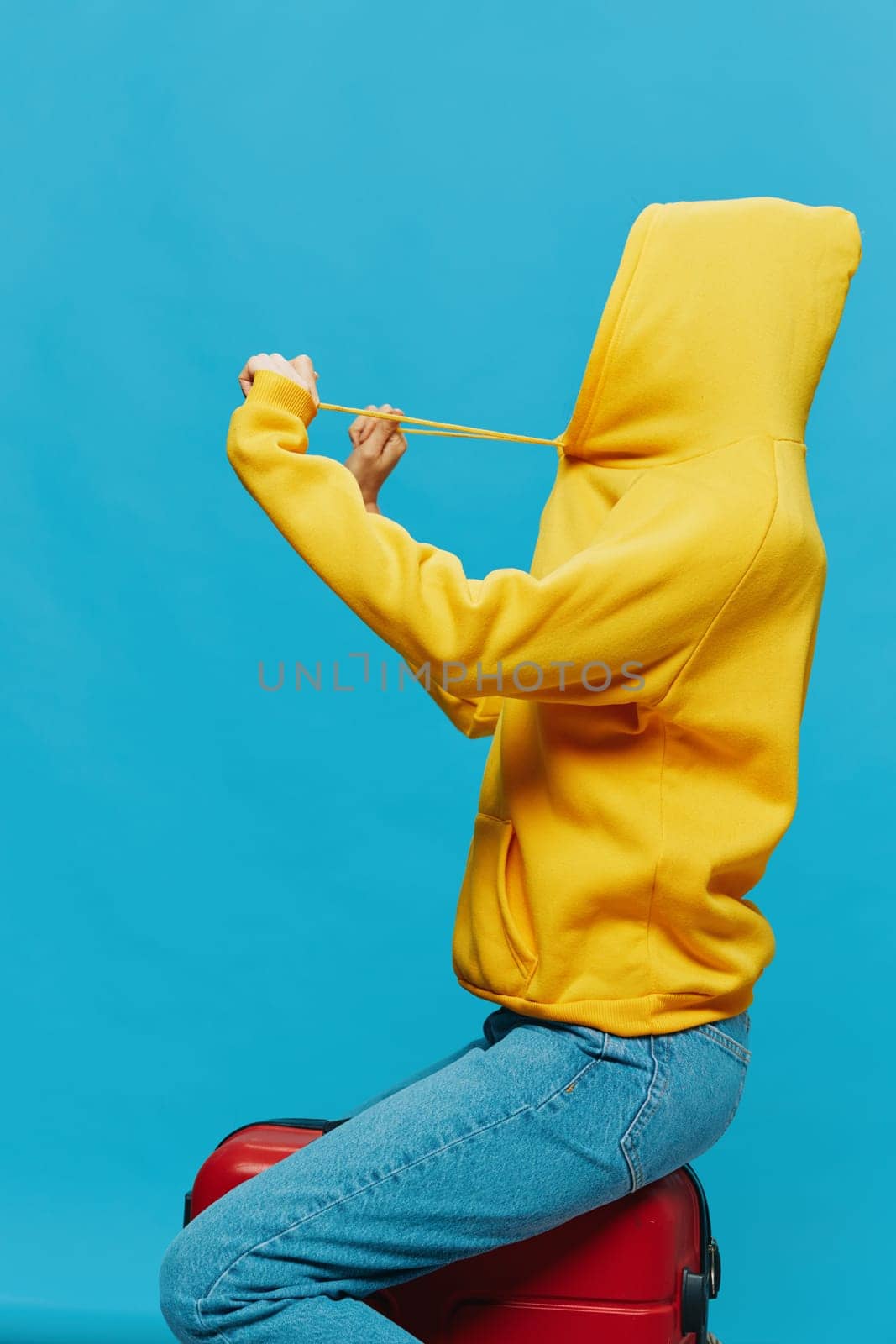 Smile woman sitting on a suitcase in a yellow hoodie, blue jeans and glasses on a blue background, packing for a trip. High quality photo