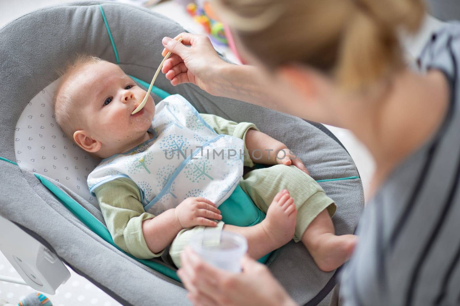 Mother spoon feeding her baby boy child in baby chair with fruit puree. Baby solid food introduction concept