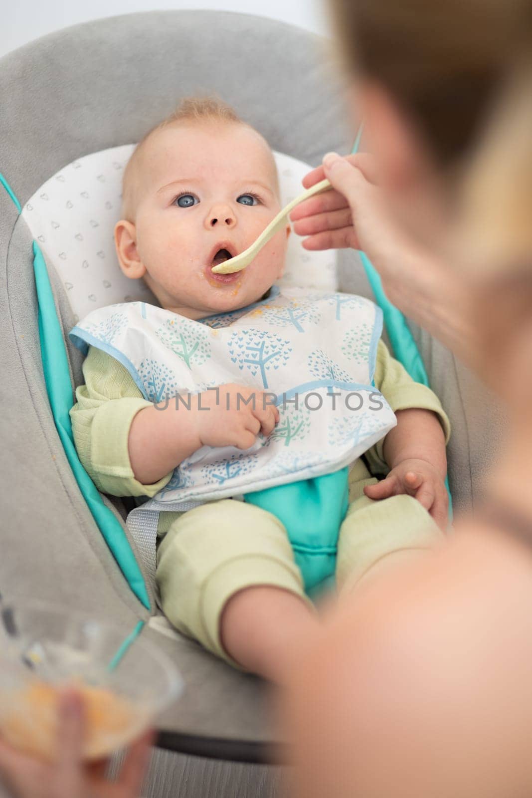 Mother spoon feeding her baby boy child in baby chair with fruit puree. Baby solid food introduction concept