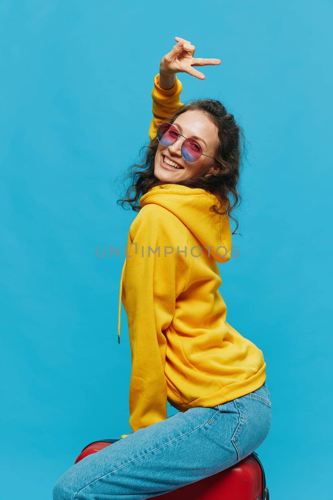 Smile woman sitting on a suitcase in a yellow hoodie, blue jeans and glasses on a blue background, packing for a trip. High quality photo