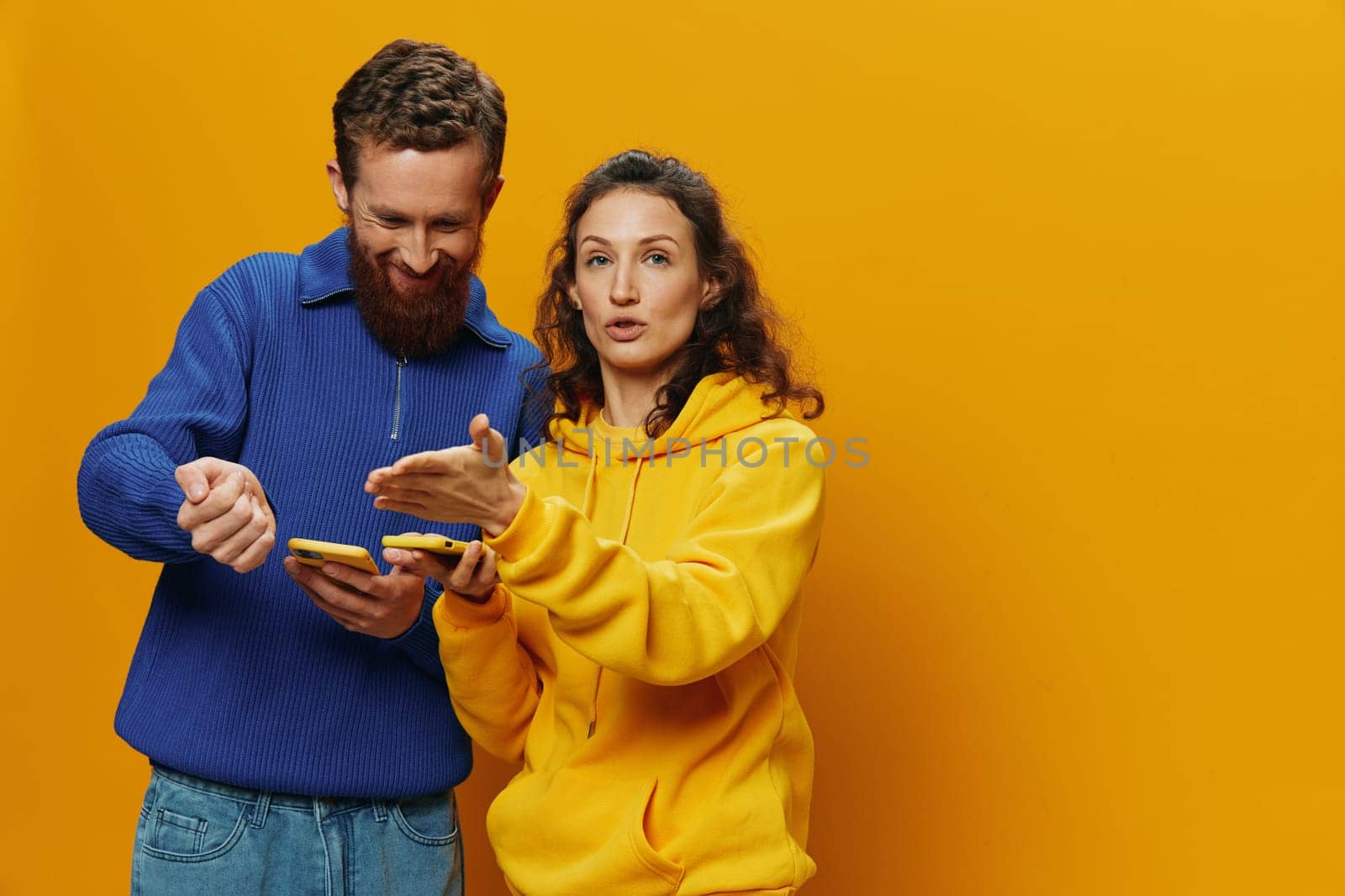 Woman man cheerful couple with phones in hand social networking and communication crooked smile fun and fight, in yellow background. The concept of real family relationships, freelancers, work online. by SHOTPRIME