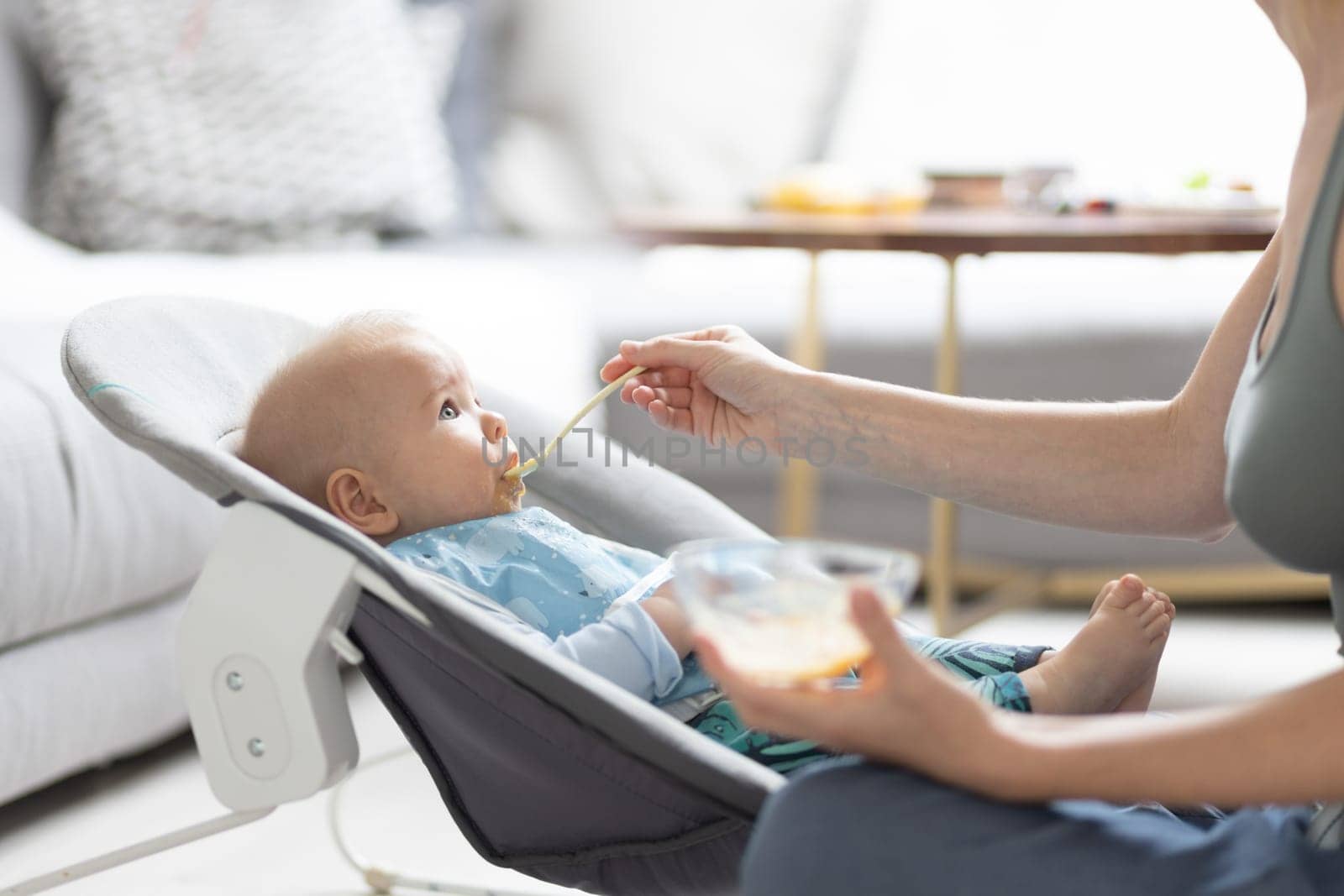 Mother spoon feeding her baby boy child in baby chair with fruit puree. Baby solid food introduction concept