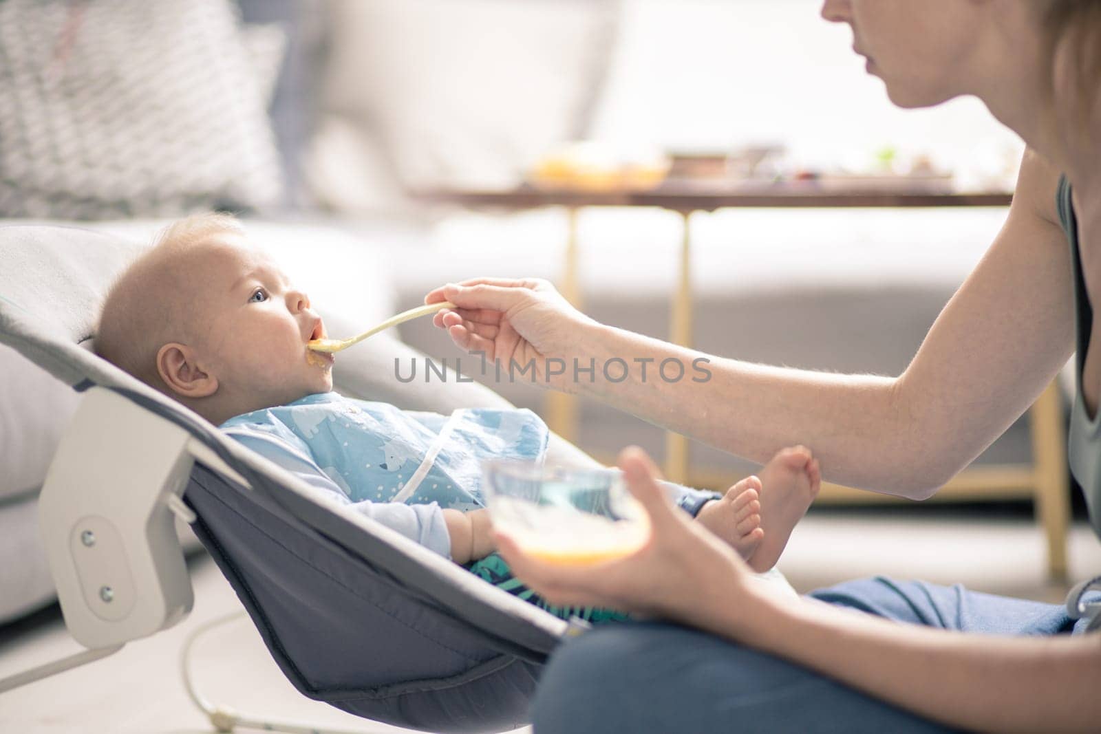 Mother spoon feeding her baby boy child in baby chair with fruit puree. Baby solid food introduction concept