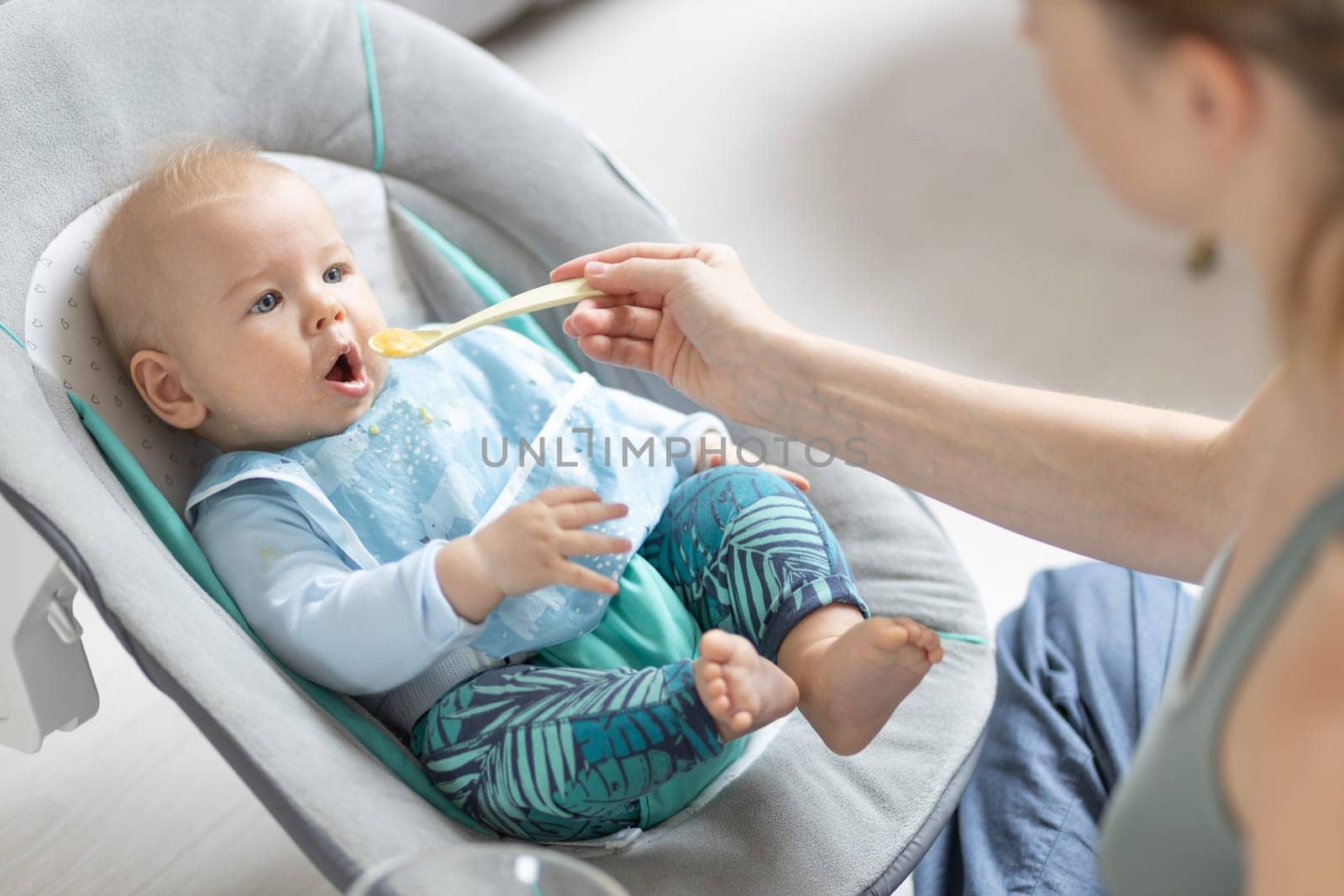 Mother spoon feeding her baby boy infant child in baby chair with fruit puree. Baby solid food introduction concept. by kasto