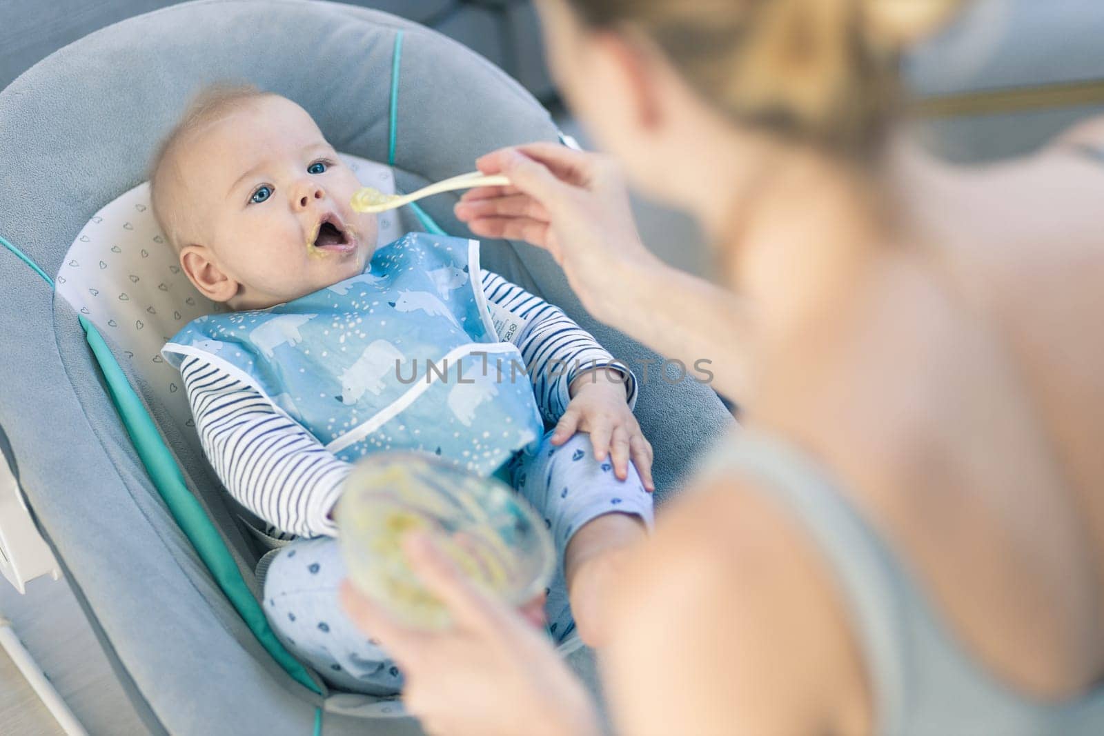 Mother spoon feeding her baby boy infant child in baby chair with fruit puree. Baby solid food introduction concept. by kasto