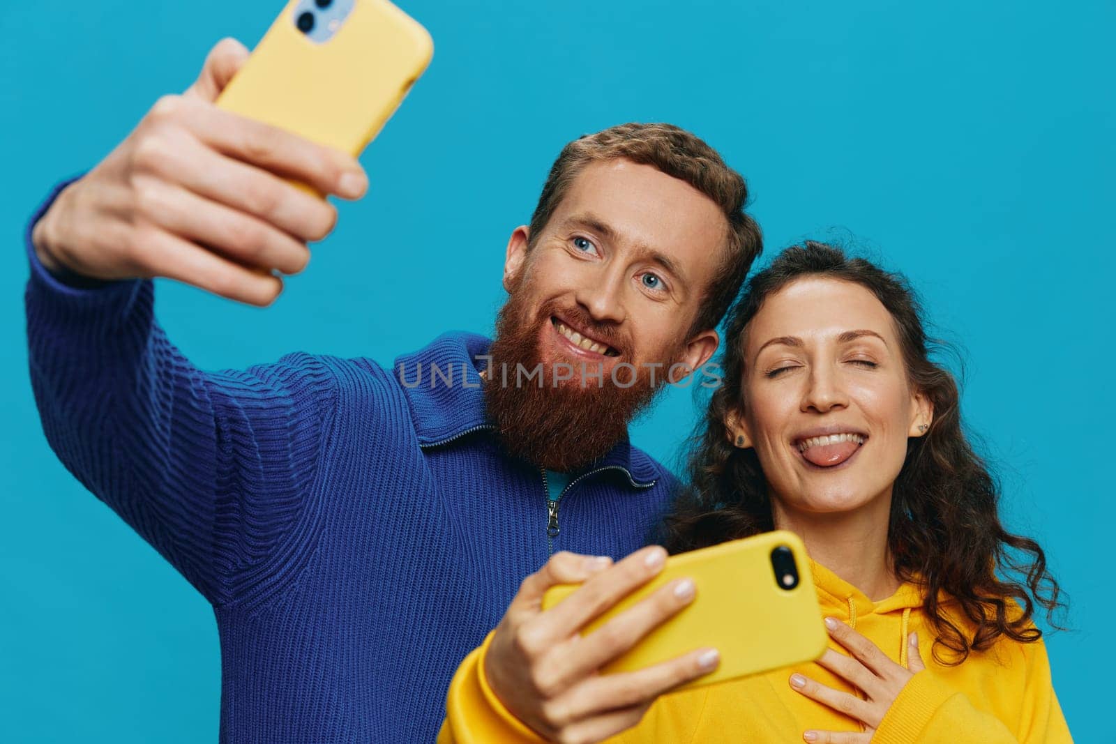 Woman and man funny couple with phones in hand taking selfies crooked smile fun, on blue background. The concept of real family relationships, talking on the phone, work online. High quality photo