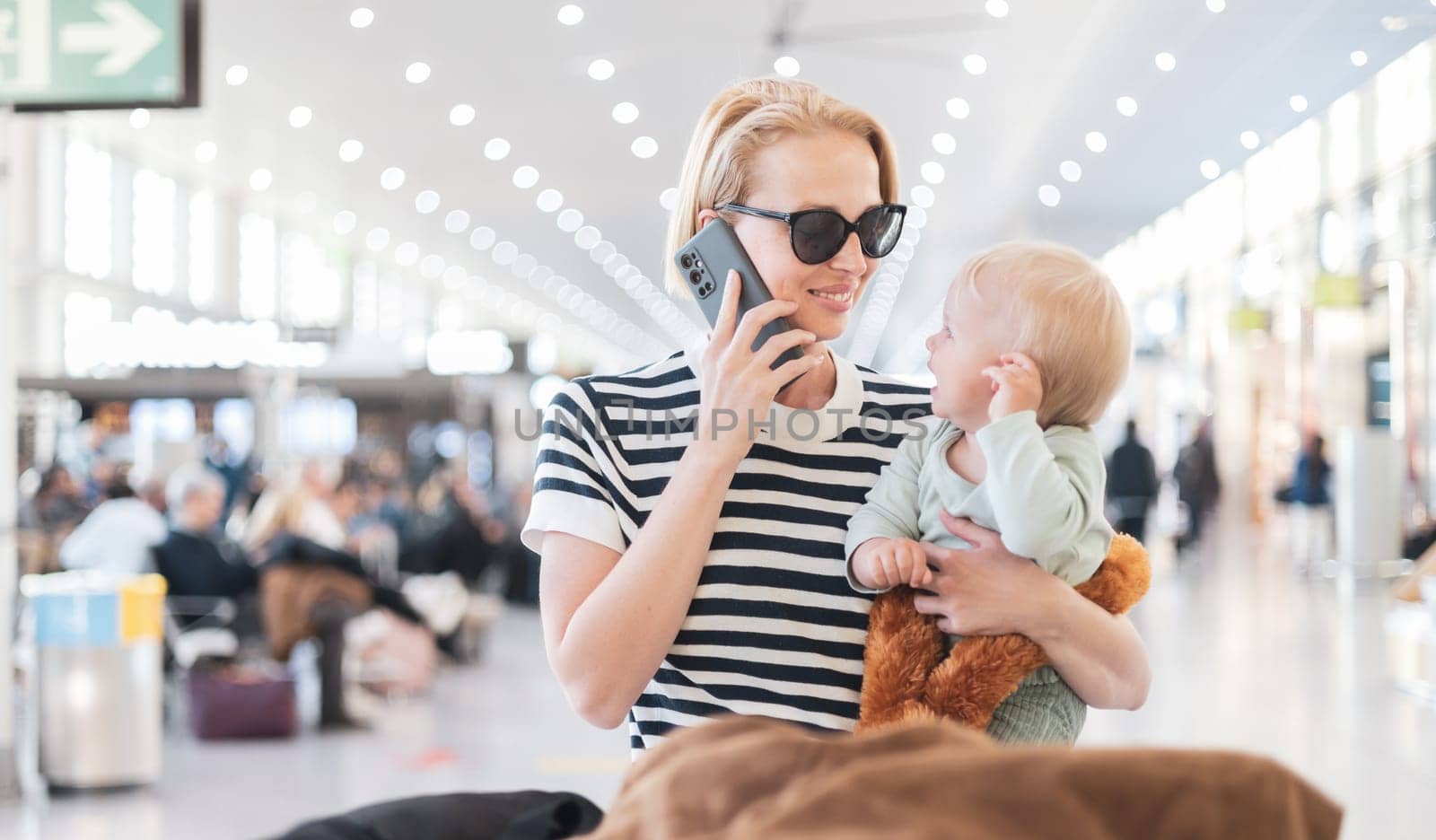 Mother talking on mobile phone while traveling with child, holding his infant baby boy at airport terminal waiting to board a plane. Travel with kids concept. by kasto