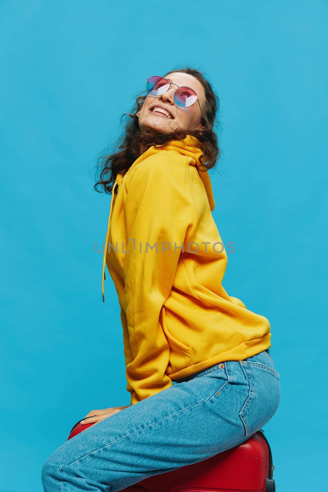 Smile woman sitting on a suitcase in a yellow hoodie, blue jeans and glasses on a blue background, packing for a trip. High quality photo