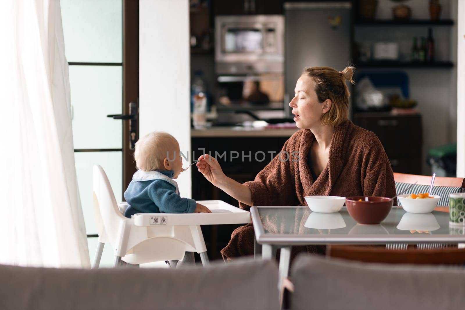 Cheerful mother wearing bathrope spoon feeding her infant baby boy child sitting in high chair at the dining table in kitchen at home in the morning. by kasto