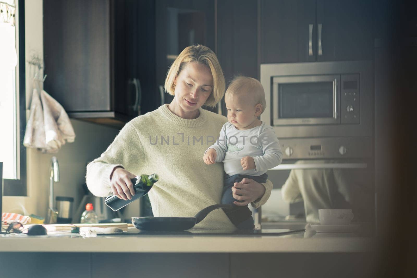 Happy mother and little infant baby boy together making pancakes for breakfast in domestic kitchen. Family, lifestyle, domestic life, food, healthy eating and people concept. by kasto