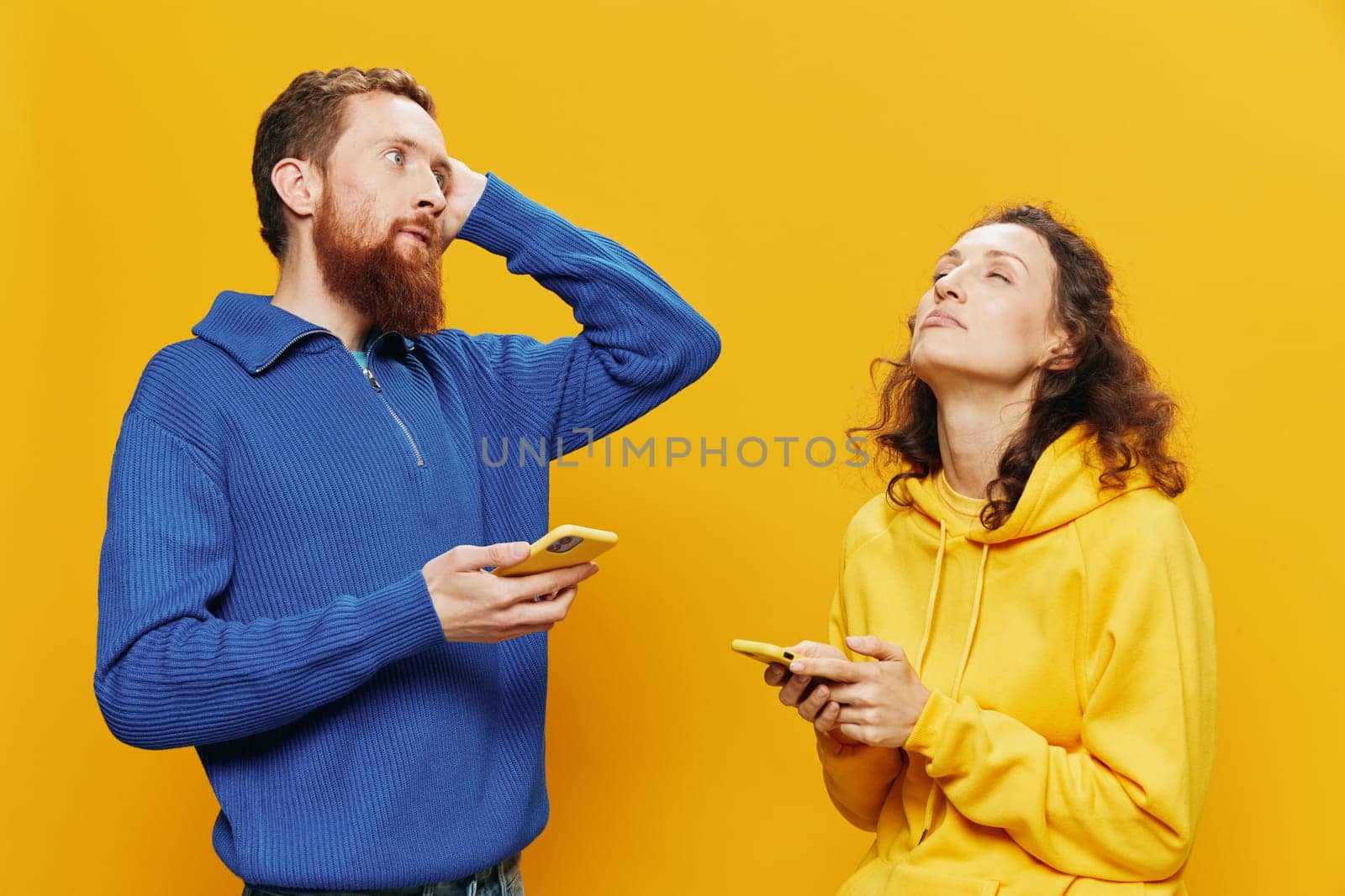 Woman man cheerful couple with phones in hand social networking and communication crooked smile fun and fight, in yellow background. The concept of real family relationships, freelancers, work online. by SHOTPRIME