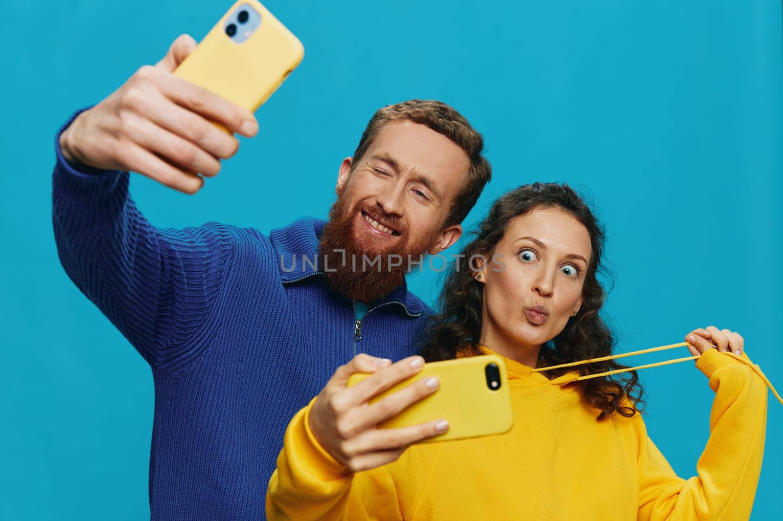 Woman and man funny couple with phones in hand taking selfies crooked smile fun, on blue background. The concept of real family relationships, talking on the phone, work online. High quality photo