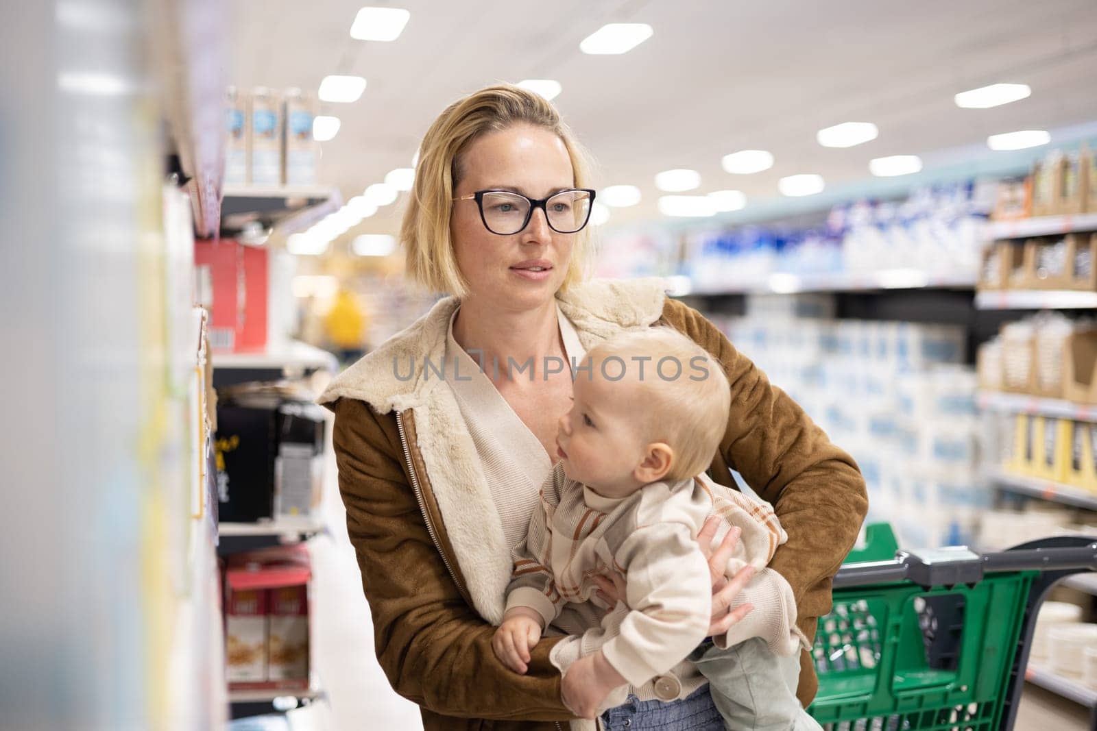 Caucasian mother shopping with her infant baby boy child choosing products in department of supermarket grocery store