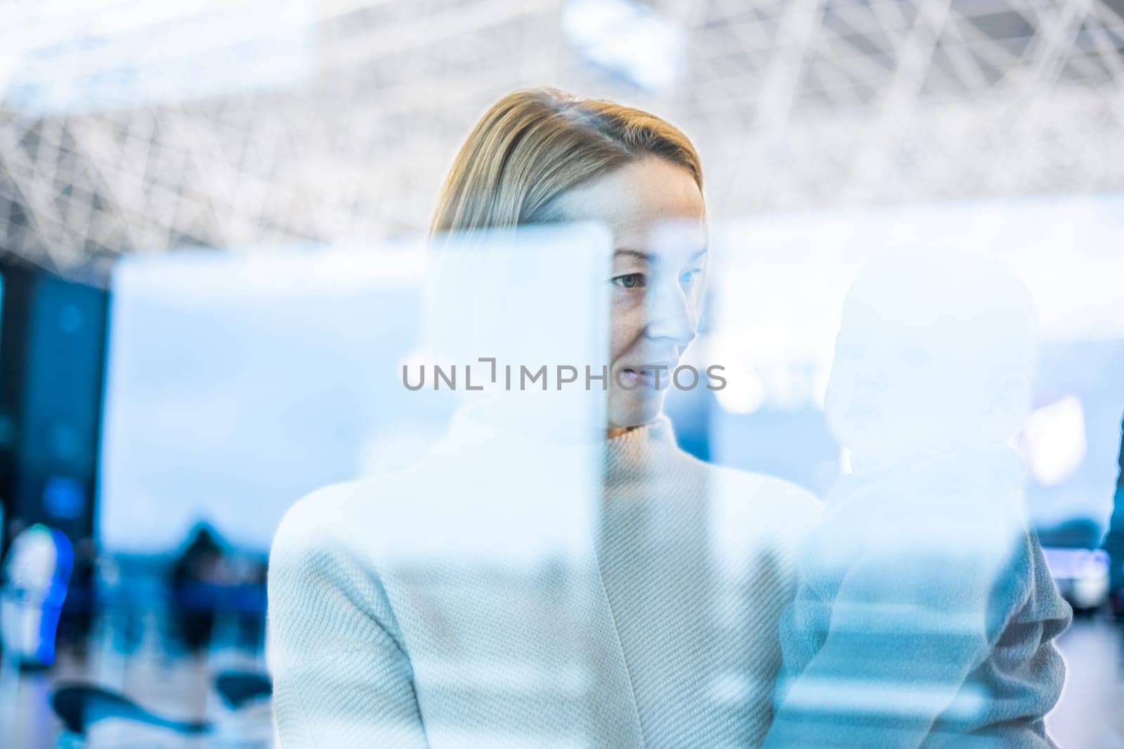 Thoughtful young mother looking trough window holding his infant baby boy child while waiting to board an airplane at airport terminal departure gates. Travel with baby concept. by kasto