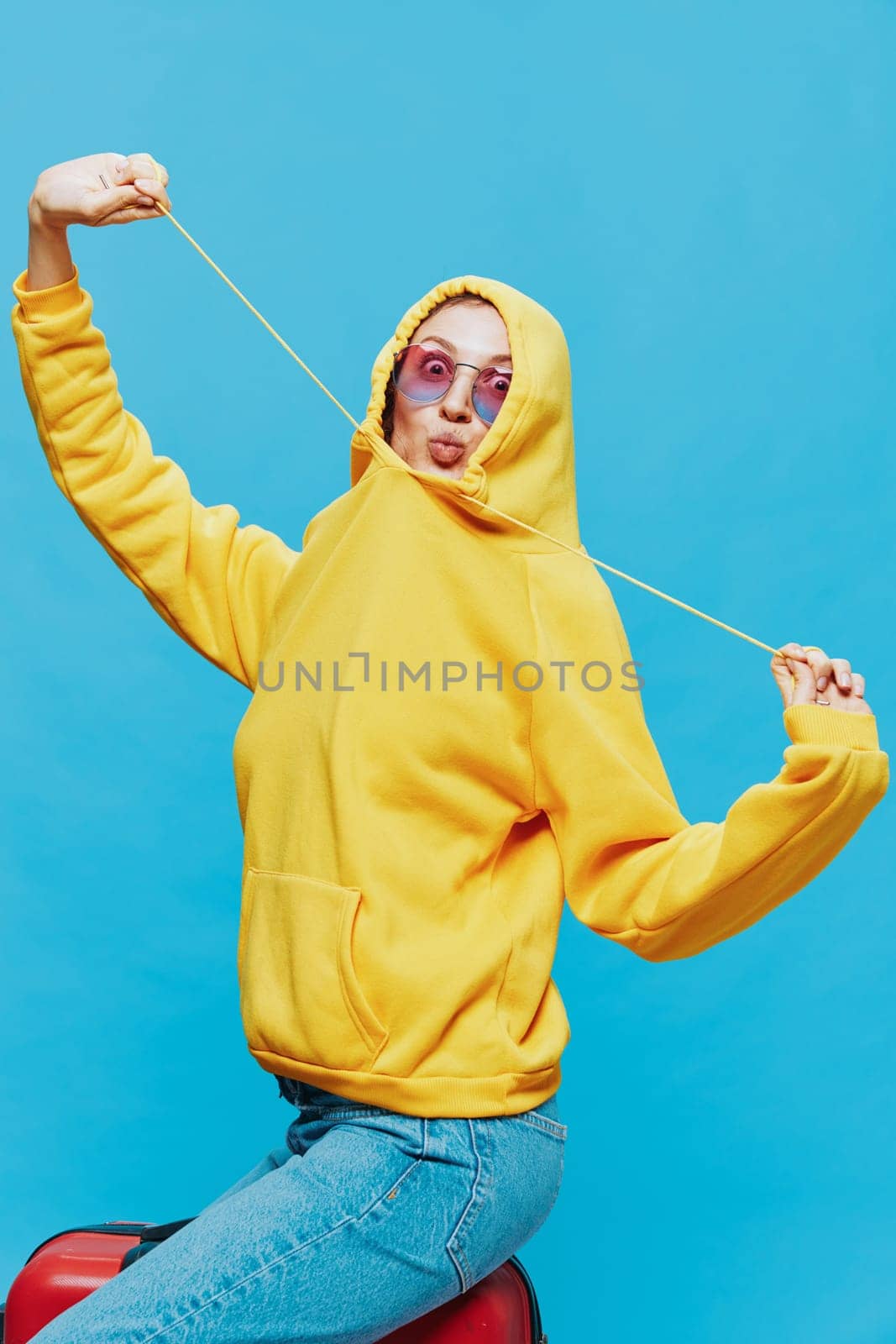 Smile woman sitting on a suitcase in a yellow hoodie, blue jeans and glasses on a blue background, packing for a trip. High quality photo