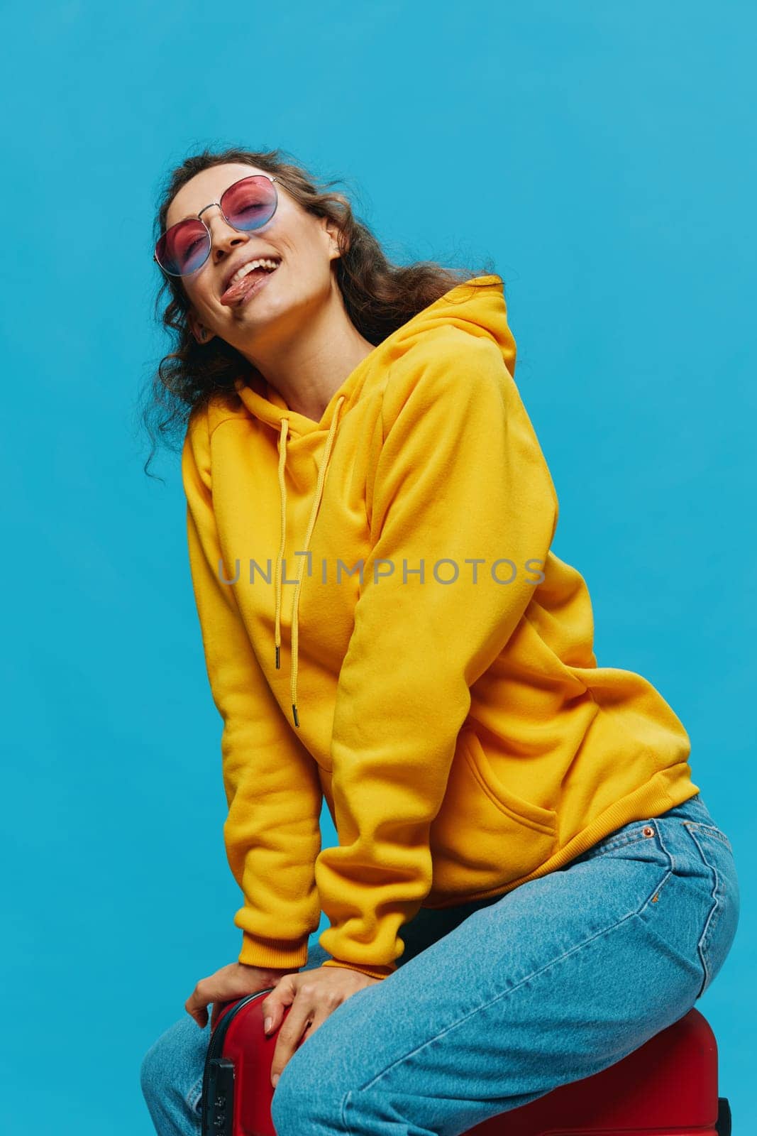 Smile woman sitting on a suitcase in a yellow hoodie, blue jeans and glasses on a blue background, packing for a trip by SHOTPRIME