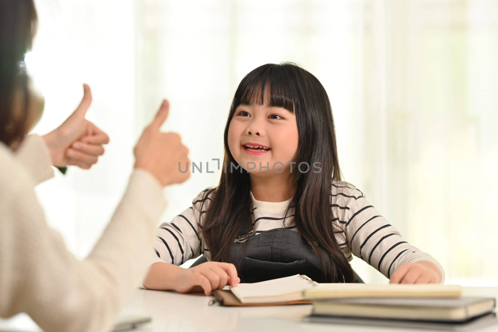 Cute little asian schoolgirl doing homework with happy mother. Distance education, homeschooling concept.