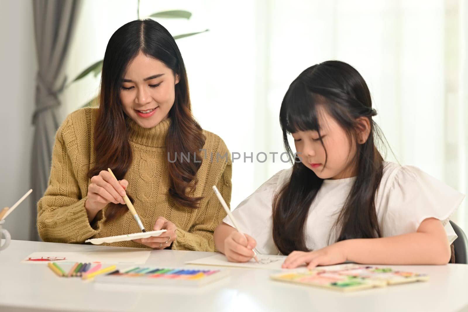 Mother and daughter drawing together in bright living room. Family home leisure concept.