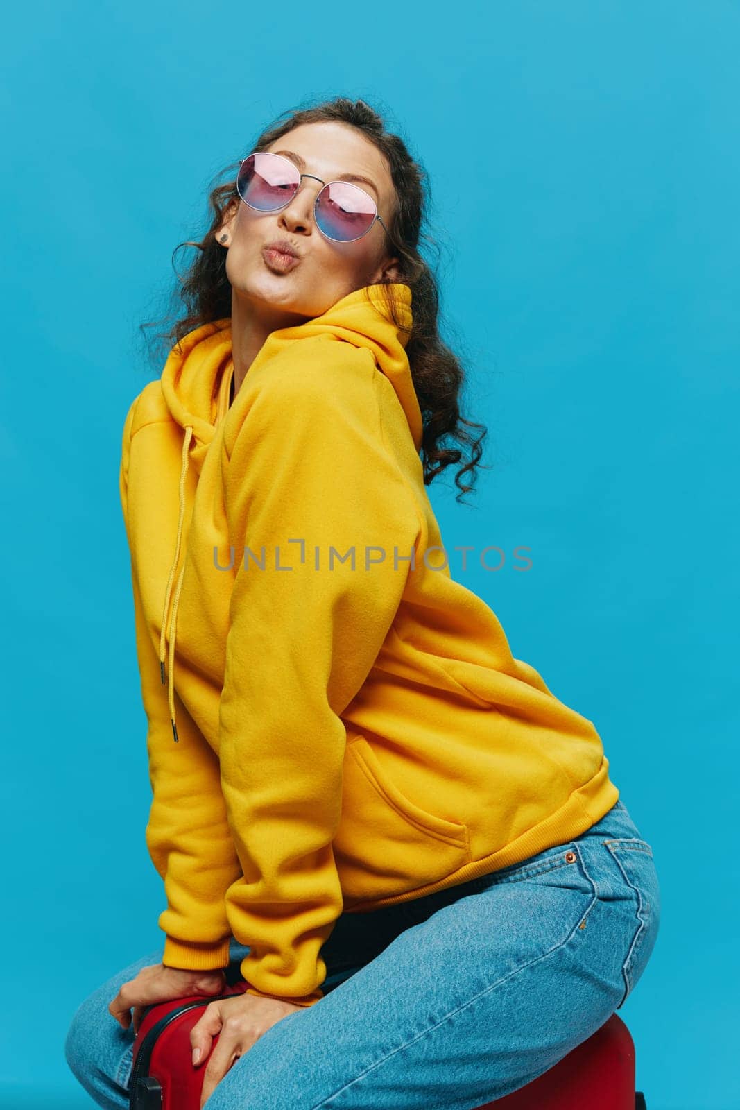 Smile woman sitting on a suitcase in a yellow hoodie, blue jeans and glasses on a blue background, packing for a trip. High quality photo