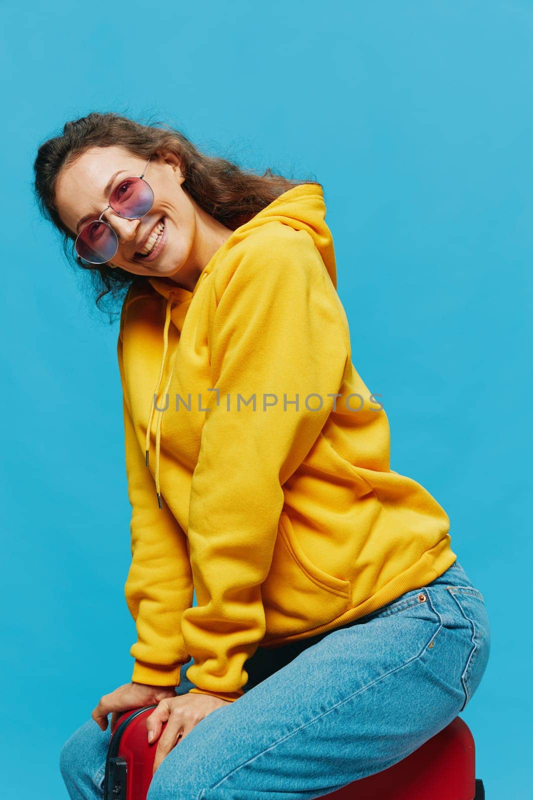 Smile woman sitting on a suitcase in a yellow hoodie, blue jeans and glasses on a blue background, packing for a trip by SHOTPRIME