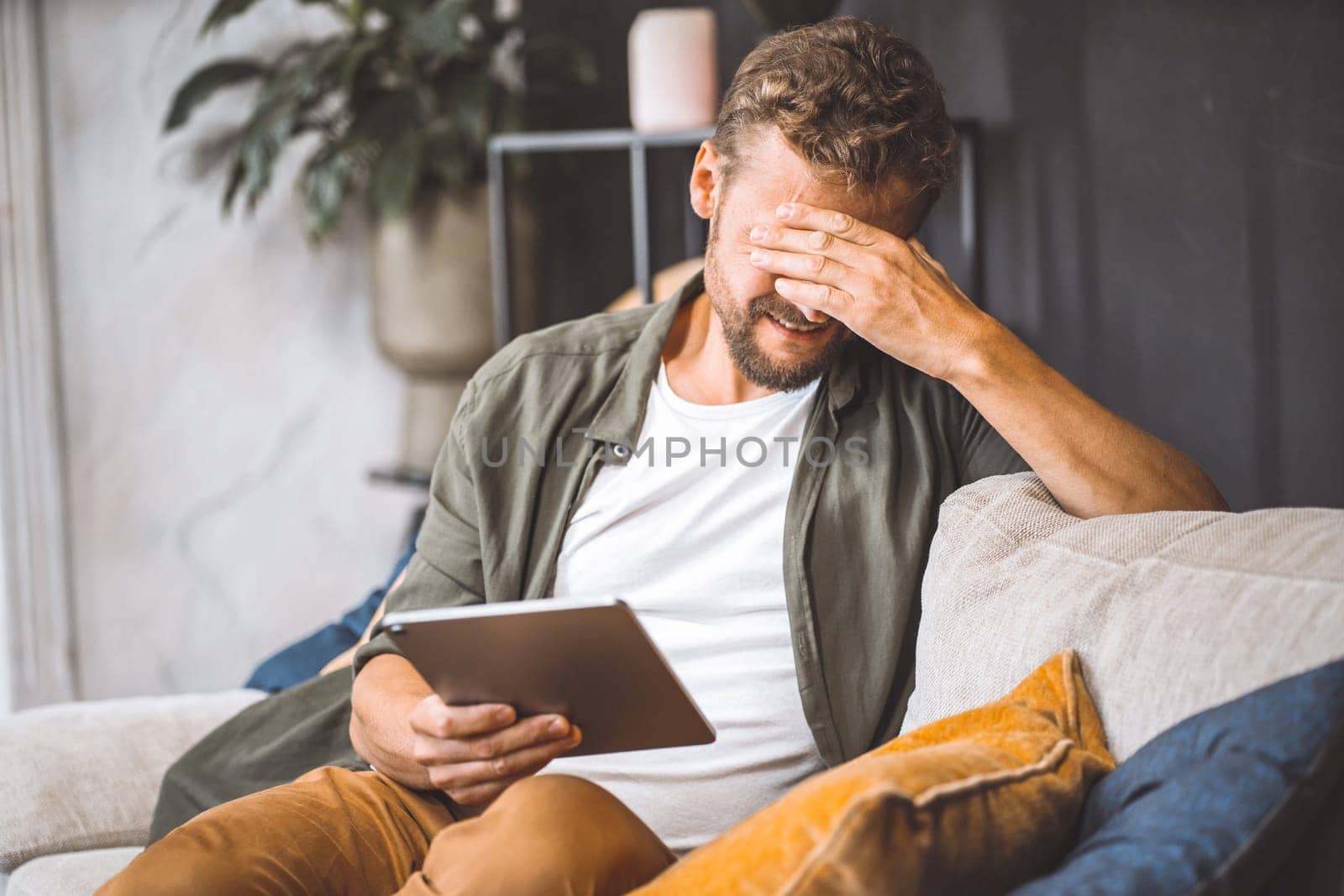 Sad man sitting on sofa home, holding tablet PC, making facepalm gesture. Frustration and disappointment on face palpable, as if something has gone wrong with technology or communication through internet. High quality photo