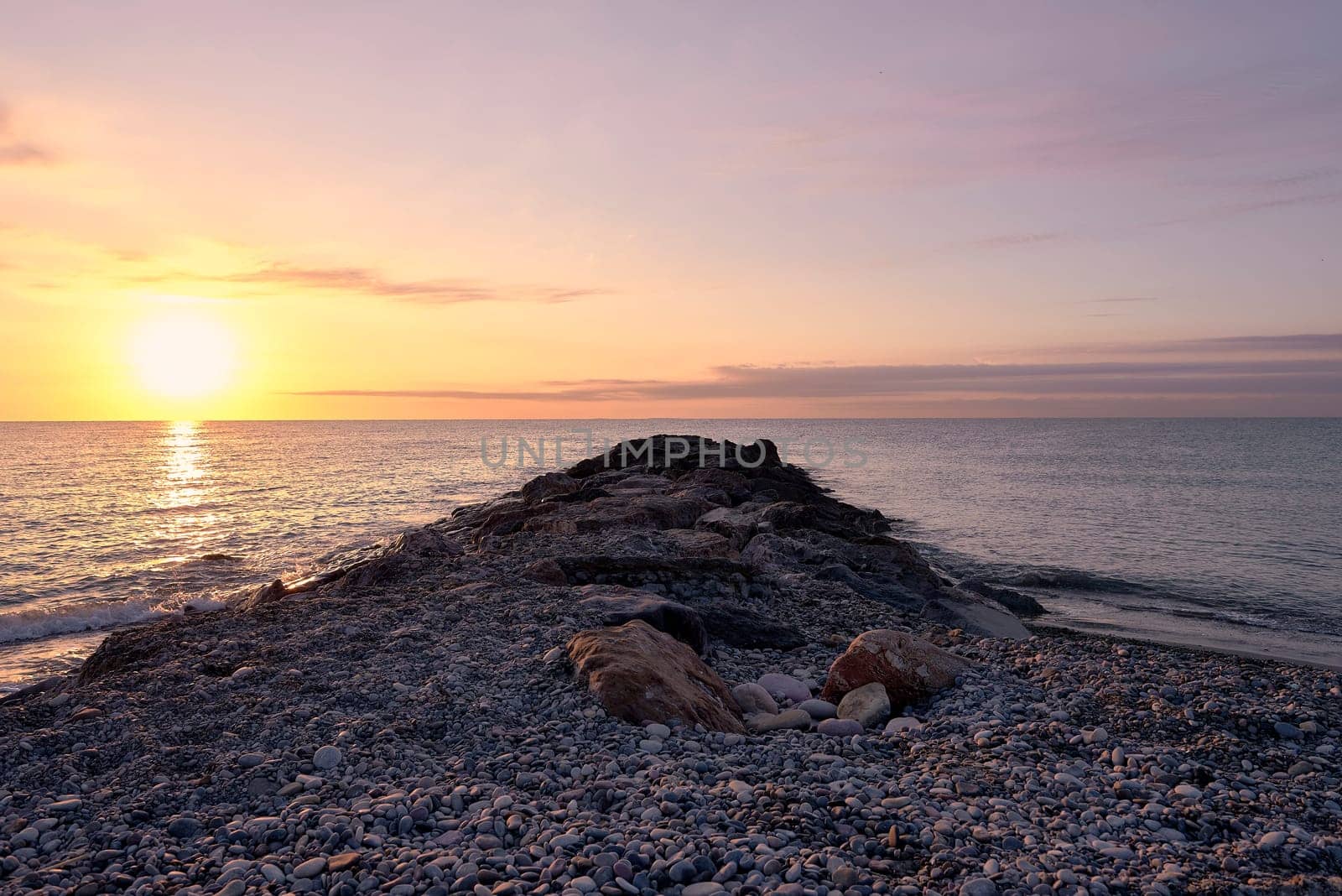 Breakwater on the rocky beach at sunrise. Lonely beach, sunshine, orange sky, small stones, calm sea