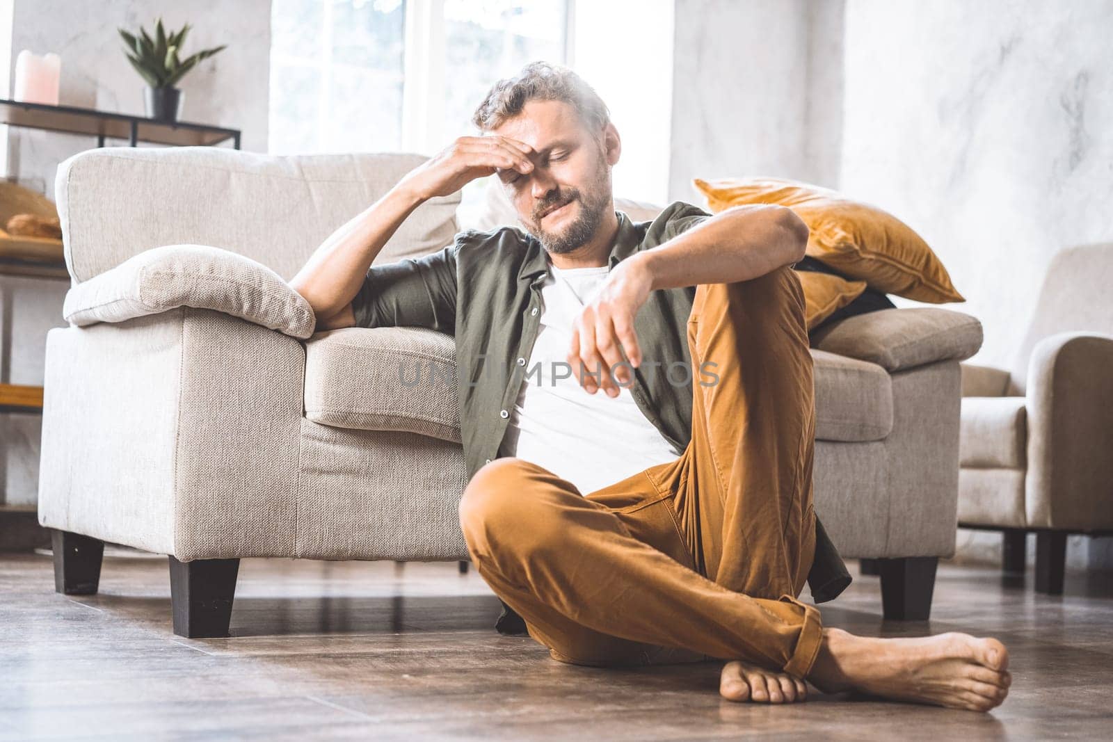 Middle-aged man resting on floor next to sofa home, moment of contemplation or crisis. Difficulties and challenges faced by men in society as they age, with focus on theme of middle age and its associated struggles. by LipikStockMedia