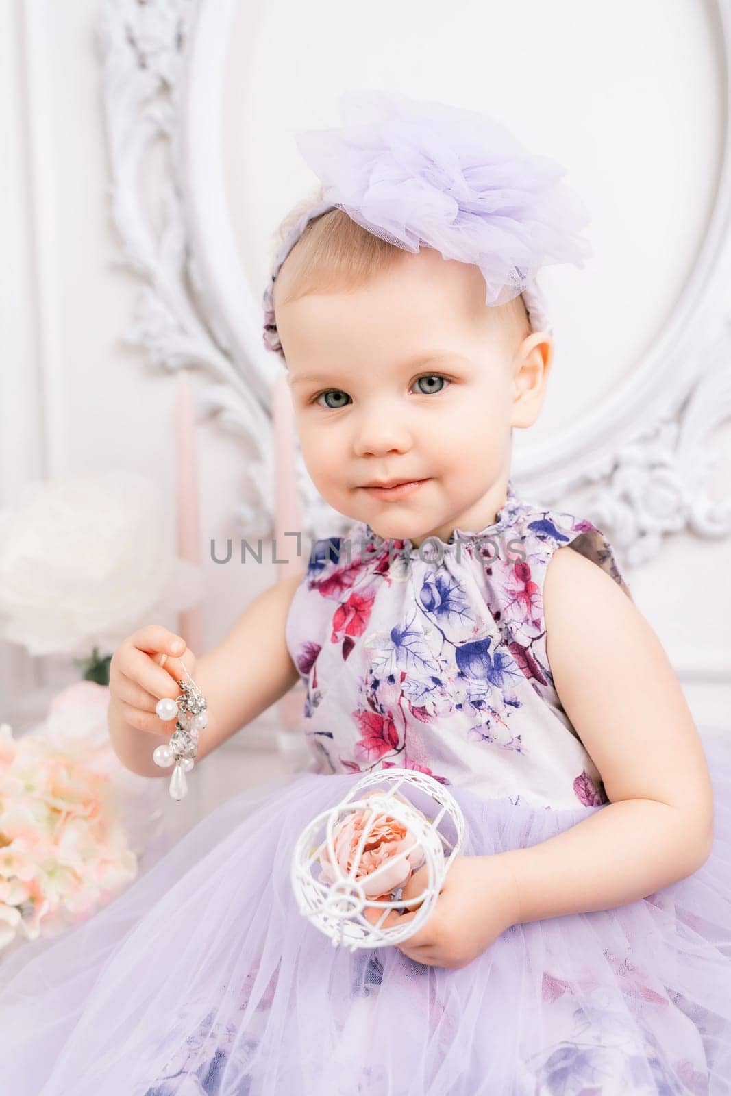 Baby girl elegant dress. A one-year-old girl in a puffy dress and a cute bow poses against the backdrop of a bright room with a dressing table and flowers