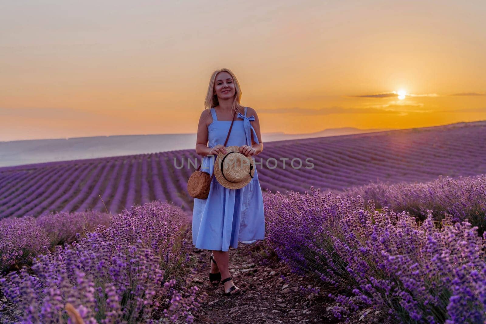 Woman lavender field sunset. Romantic woman walks through the lavender fields. illuminated by sunset sunlight. She is wearing a blue dress with a hat