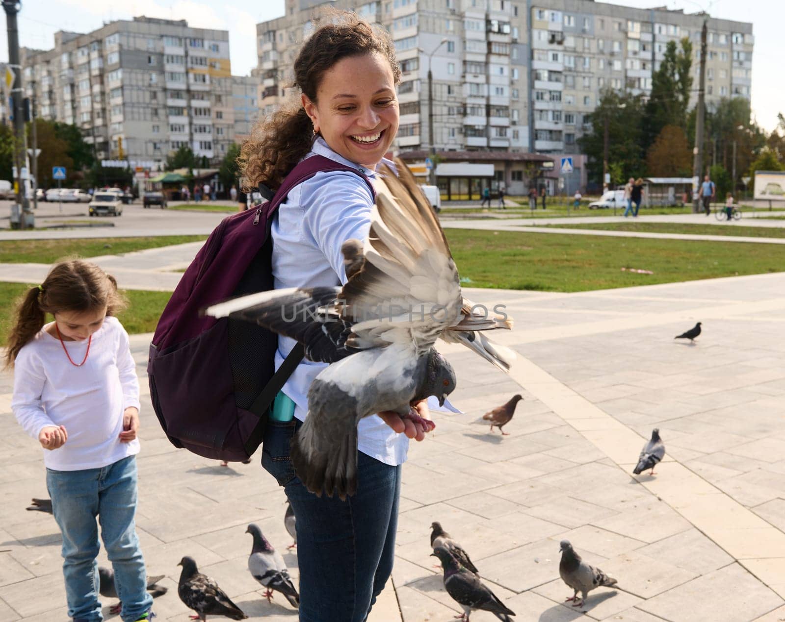 Smiling multi ethnic woman tourist with backpack, expressing positive emotions while feeding rock pigeons and feral doves in the urban square. The concept of people, nature and kindness for animals
