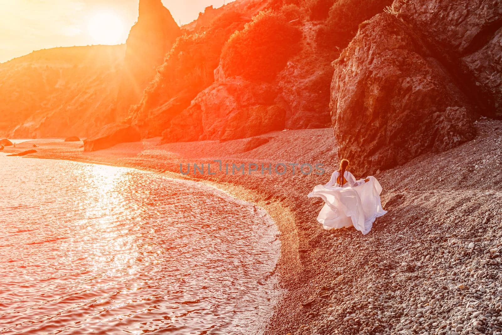 Mysterious woman silhouette long hair walks on the beach ocean water, sea nymph wind listens to the wave. Throws up a long white dress, a divine sunset. Artistic photo from the back without a face by Matiunina