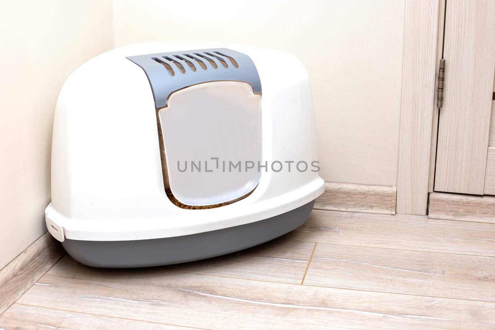 A large, gray, triangular plastic, enclosed litter box for a cat sits on the floor in the corner of a bright room.