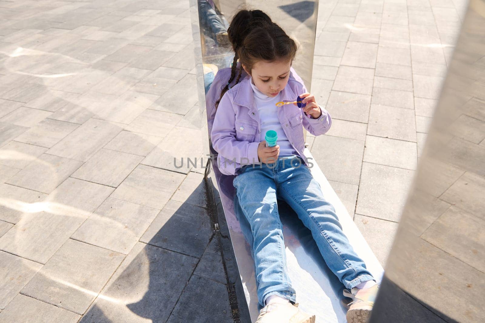 View from above of Caucasian adorable little child girl sitting on metal mirror bench, blowing soap bubbles while resting in urban park on a sunny day. Leisure activity. Happy carefree childhood