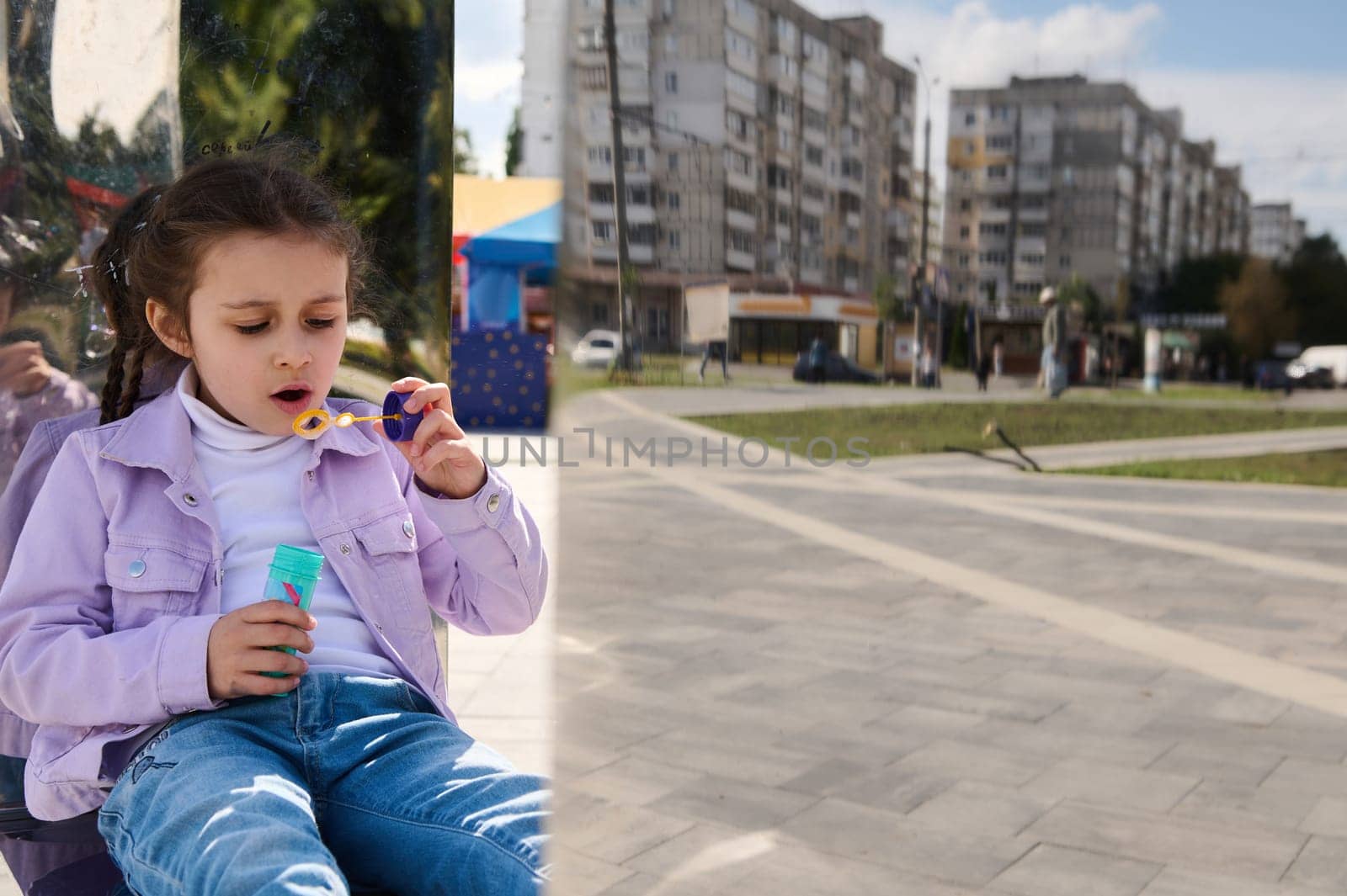 Reflection in a metal mirror bench of a charming girl blowing soap bubbles in a city park by artgf