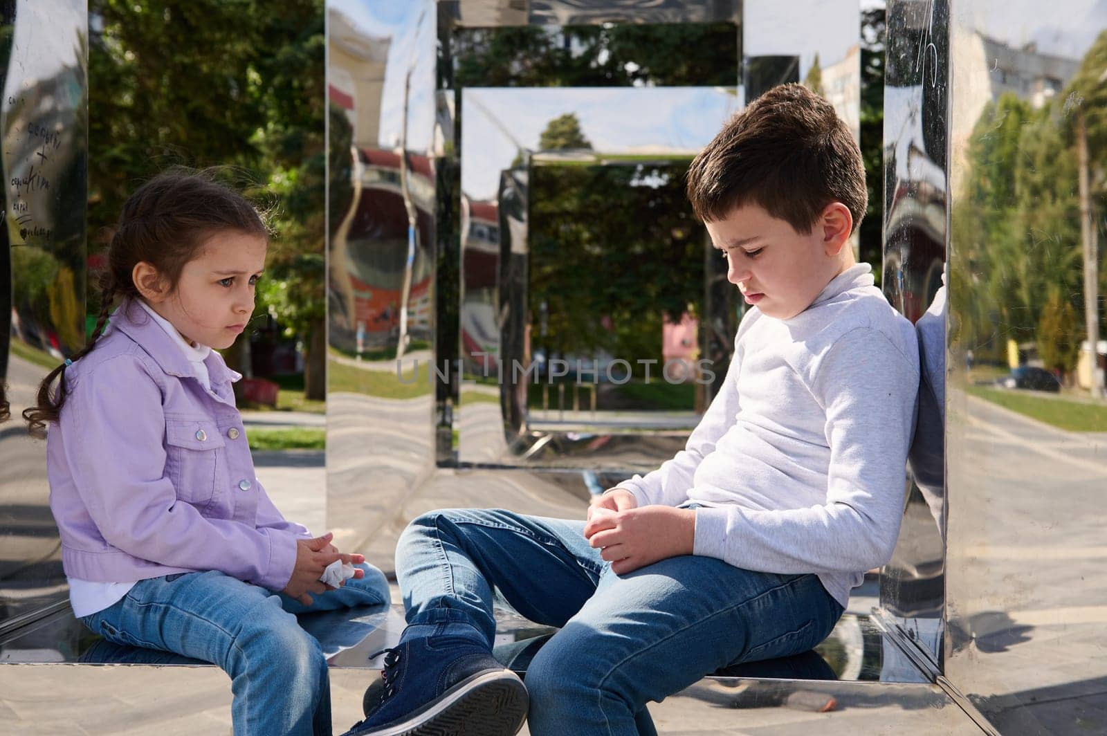 Portrait of two adorable kids boy and girl, brother and sister sitting on mirror bench in urban park, looking sad during family outing. Friendships and human relationships. People. Lifestyle. Leisures